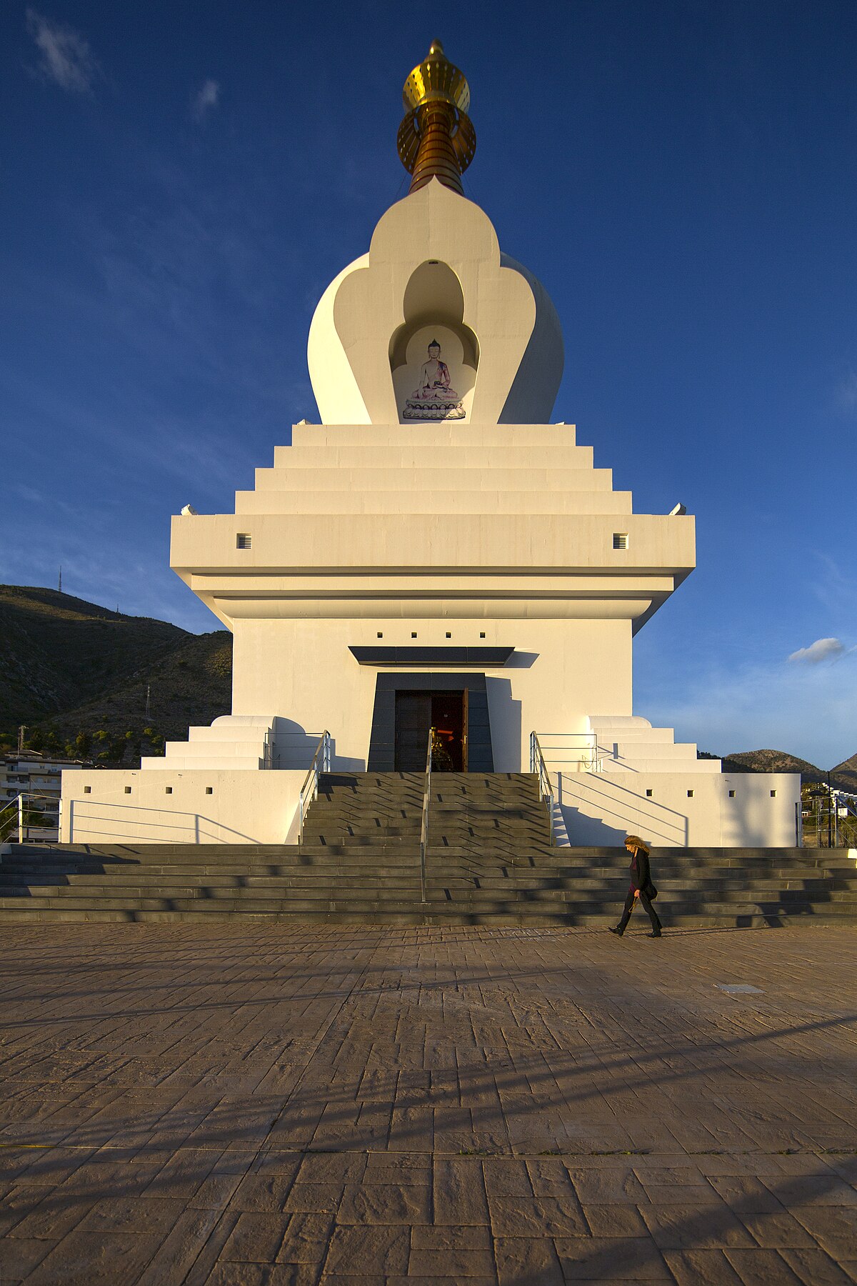 Alt text: White stupa with golden spire, steps leading up, clear blue sky, mountainous background.